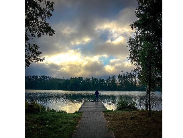 mężczyzna trzyma wędkę stojąc na pomoście nad jeziorem do pomostu prowadzi ścieżka brukowa