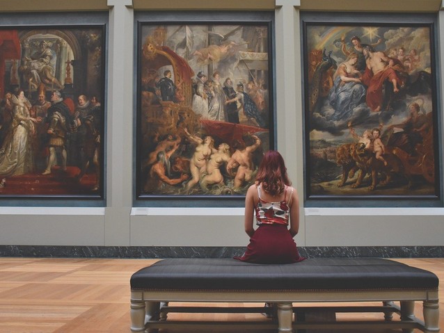 Młoda kobieta siedzi na ławce w sali muzealnej przed trzema dużymi obrazami wiszącym na ścianie przed nią