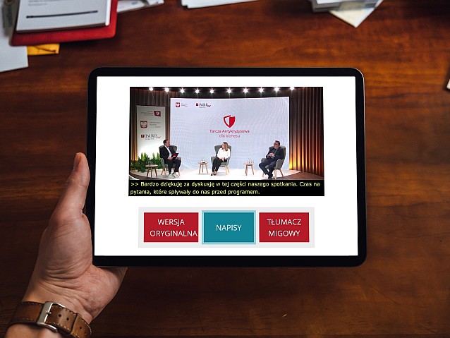 Dłoń trzyma tablet, na którego ekranie jest wyświetlana debata nt. Tarczy Antykryzysowej z udziałem 3 osób. Pod wideo są napisy z treścią tego, co mówią, a niżej klawisze do wyboru: wersja oryginalna, napisy, tłumacz migowy