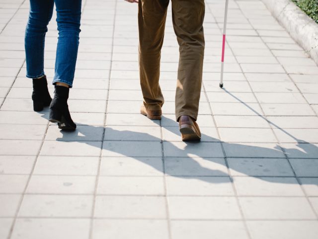Kobieta i mężczyzna idą chodnikiem ułożonym z płyt. Mężczyzna używa białej laski