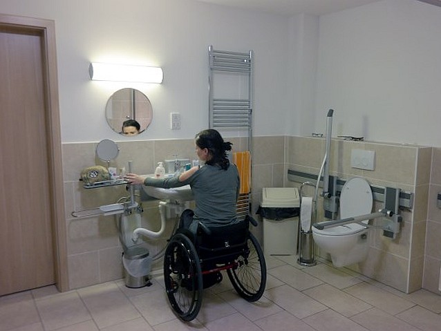 Kobieta na wózku przy umywalce w przestronnej dostosowanej toalecie. Po prawej miska ustępowa z poręczami
