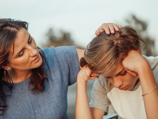 kobieta kładzie rękę na głowie nastolatka który siedzi smutny z głową opartą na rękach