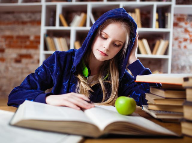 dziewczyna w bluzie z kapturem na głowie siedzi przy stole nad otwartymi książkami przed nią zielone jabłko