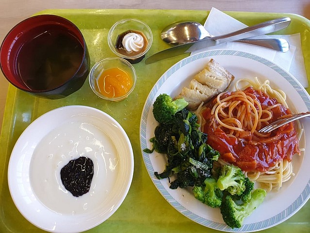 Taca z różnego rodzaju jedzeniem, w tym: spaghetti z sosem, brokuły, ryba, budynie, coś do picia