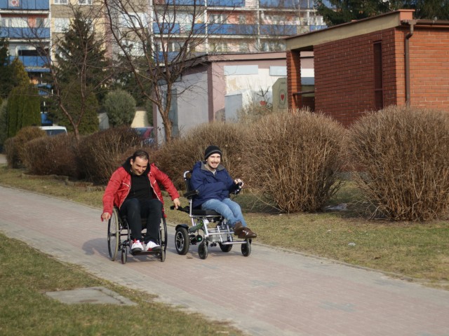 Michał Koterski jedzie wózkiem, obok niego mężczyzna poruszający się na mózgu. Jadą chodnikiem pod blokiem