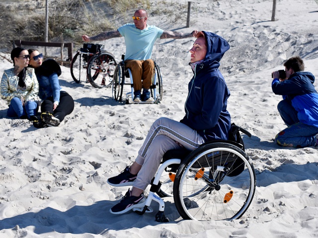 kobieta na wózku i mężczyzna na wózku na drugim planie pozują do zdjęć na plaży