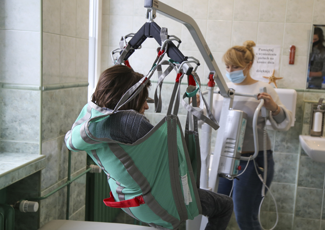 kobieta z niepełnosprawnością jest przenoszona na specjalnym urządzeniu, sterowanym przez kobietę w maseczce