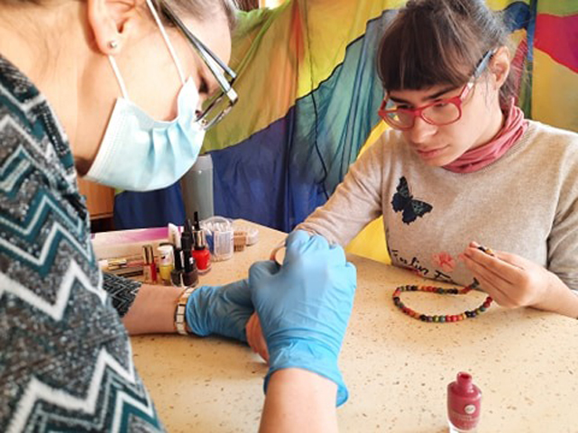 kobieta w maseczce i rękawiczkach maluje paznokcie młodej kobiecie z niepełnosprawnością