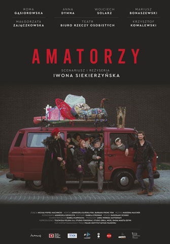 Plakat filmu pt. Amatorzy. W czerwonym busie otwarte są drzwi. Wyglądają z niego osoby z zespołem Downa. Obok busa stoi dwóch mężczyzn. Na busie są doczepione bagaże