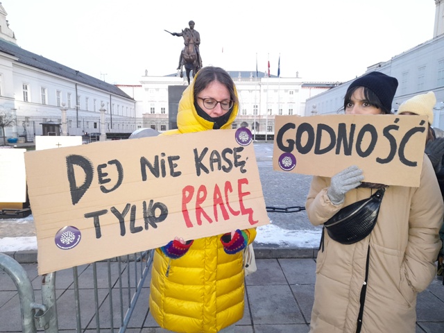 Dwie kobiety stoją pod Pałacem prezydenckim z transparentmai. Na jednym jest napisane: "Daj nie kasę, tylko pracę" a na drugim: godność. 