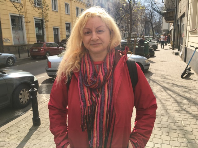 Sofia Markina - kobieta w średnim wieku, okrągła twarz, długie falowane blond włosy spoczywają na ramieniu, ma czerwoną sportową kurtkę, ręce trzyma w kieszeniach. Delikatnie się uśmiecha