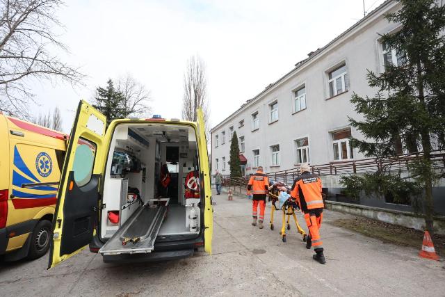 ratownicy wiozą na łóżku osobę z niepełnosprawnością. Po prawej jest budynek, po ich lewej karetka