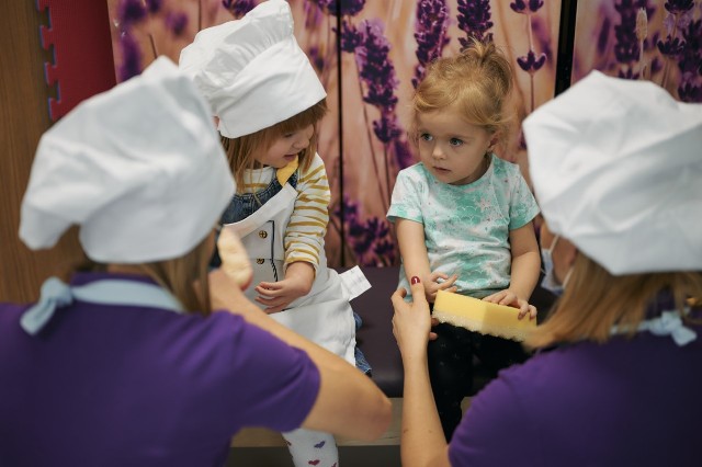 dwie młode kobiety w czepkach kucharskich kucają nad dwiema siedzącymi dziewczynkami z niewykształconymi rękami, panie zachęcają do wspólnego gotowania