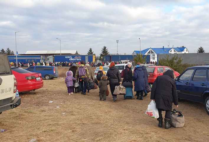 Ukraińscy uchodźcy idą drogą polną, gdzie stoją puste auta w kierunku drogi asfaltowej, gdzie są tiry, wśród których są tłumy ludzi