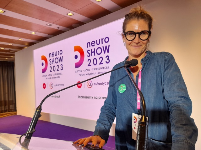 Lotta Borg Skoglund, szczupla blondynka w dżinsowej koszuli stoi przy pulpicie na scenie. Za nią na ekranie wyświetlane są dwa loga konferencji NeuroShow 2023