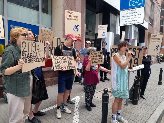 Przed wejsciem do gmachu Ministerstwa Rodziny i Polityki Społecznej stoją uczestnicy protestu z transparentami. Na transparentach napisano m.in. 2119 szablą odbierzemy (napisowi towarzyszy rysunek szabelki),Dlaczego politycy "za życiem" nie dają żyć, Nic nas nie zagłuszy, nic nas nie powstrzyma, godne życie nie tylko dla bogaczy, Stop przemocy finansowej i Protest 2119. Przed protestującymi stoi Karolina Kosecka z mikrofonem. 