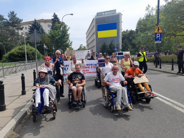 Ulicą Wiejską idzie duża grupa protestujących osób z niepełnosprawnością i ich opiekunów. Wśród nich jest Iwona Hartwich i jej syn. 