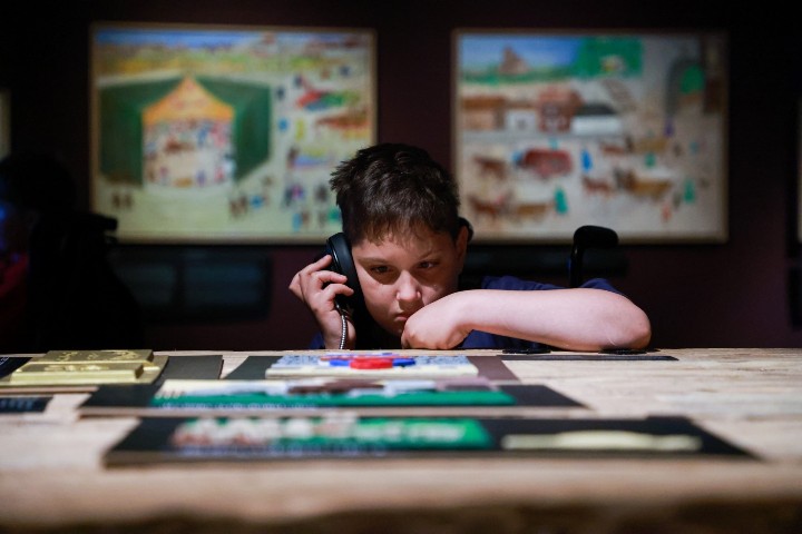 Wystawa w muzeum POLIN. Chłopiec spogląda na stół z eksponatami. Chłopiec ma założone słuchawki. Za nim znajdują się obrazy.