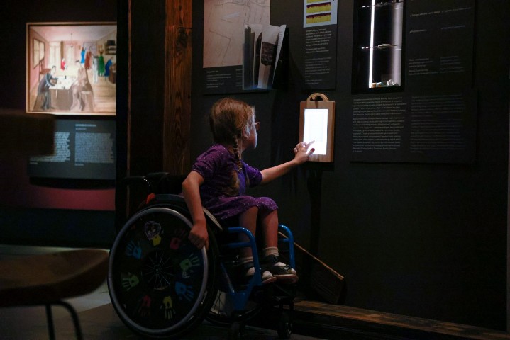 Wystawa w muzeum POLIN. Dziewczynka na wózku kilka cos na tablecie przyczepionym do ściany. Tablet jest na jej wysokości. 