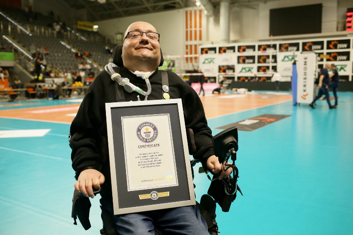Na zdjęciu znajduje się bohater artykułu - Janusz Świtaj. Mężczyzna jest rekordzistą Guinnessa w kategorii: najdłuższe życie na wentylacji mechanicznej. Na zdjęciu jest uśmiechnięty a na kolanach leży certyfikat rekordu Guinnessa