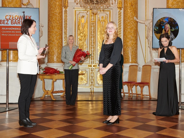 Na środku sceny podczas Gali konkursu Człowiek bez barier, jest Julia Kazimierczak, ubrana w czarną sukienkę młoda dziewczyna w okularach po lewej jest ubrana w białą marynarkę kobieta niosąca statuetkę dla Julii, po prawej Monika Kapinos. W tle jest kobieta trzymająca kwiaty.