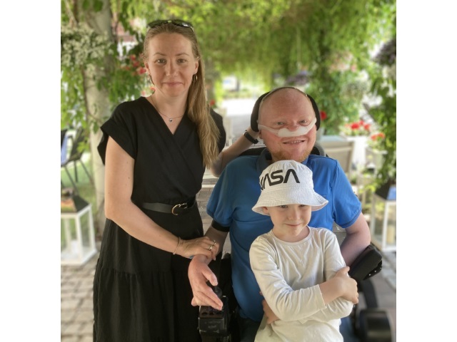 Łukasz Krasoń, młody meżczyzna na wózku elektrycznym i respiratorem pod nosem stoi obok żony, która trzyma go zarękę. Na kolanach ma dziecko. W tle ogród.