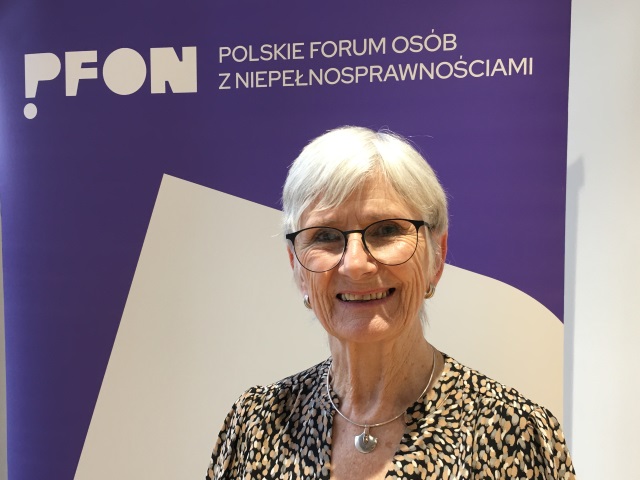 Maureen Piggot, szczupła kobieta w średnim wieku stoi na tle banera Polskiego Forum Osób z Niepełnosprawnościami.