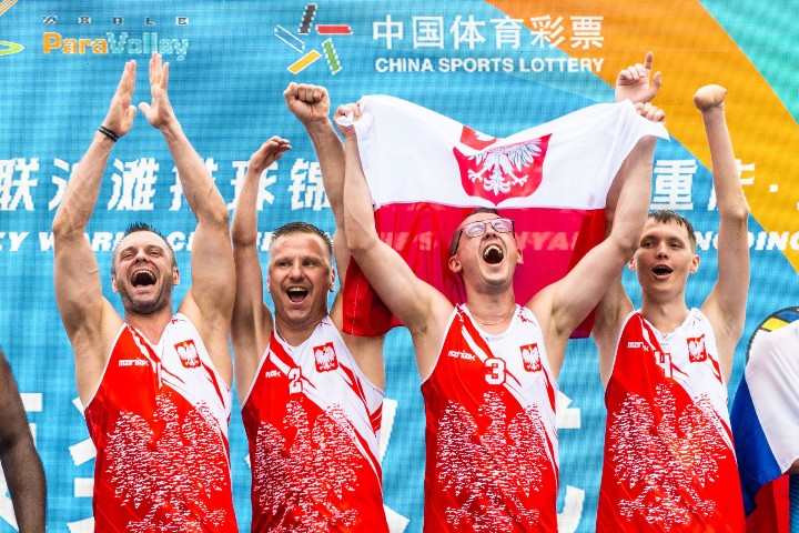 Reprezentacja polski w siatkówce plażowej. Czterech mężczyzn w biało-czerwonych koszulkach. Uśmiechają się, mają triumfalne pozy, klaszczą. Jeden z nich trzyma biało-czerwoną flagę.