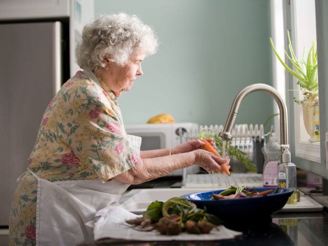 starsza pani w fartuchu myje warzywa w kuchni pod kranem