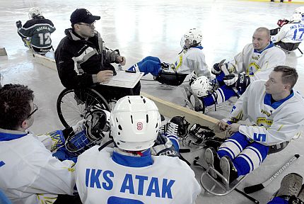 Trener siedzący na wózku instruuje grupę hokeistów na sledżach.