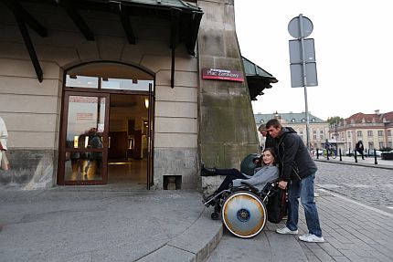 Kobieta siedzi na wózku, unoszonym przez mężczyznę, żeby pokonać wysoki krawężnik